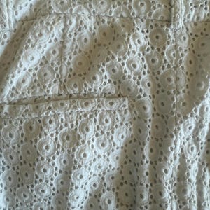 May include: Close-up of white eyelet shorts. The shorts feature a detailed, openwork design with circular patterns. The shorts have a waistband and a pocket detail, showcasing the intricate fabric texture.