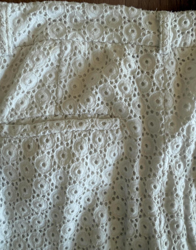 May include: Close-up of white eyelet shorts. The shorts feature a detailed, openwork design with circular patterns. The shorts have a waistband and a pocket detail, showcasing the intricate fabric texture.