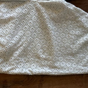 May include: Close-up of white eyelet fabric featuring a repeating circular design. The fabric has a delicate, textured appearance, with small holes creating a lace-like effect. The material is likely cotton or a similar lightweight textile.