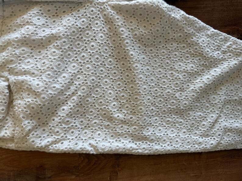 May include: Close-up of white eyelet fabric featuring a repeating circular design. The fabric has a delicate, textured appearance, with small holes creating a lace-like effect. The material is likely cotton or a similar lightweight textile.