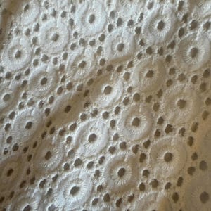 May include: Close-up of white eyelet fabric. The textile displays a repeating pattern of embroidered circles with small, evenly spaced holes. The texture is soft and the colour is a bright white, suggesting a delicate textile.