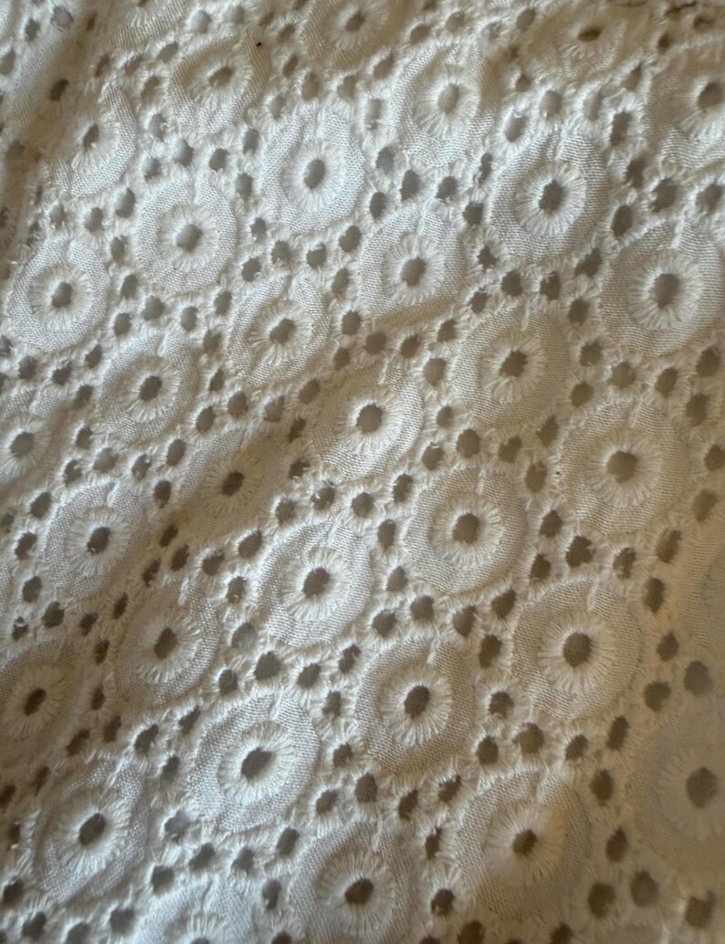May include: Close-up of white eyelet fabric. The textile displays a repeating pattern of embroidered circles with small, evenly spaced holes. The texture is soft and the colour is a bright white, suggesting a delicate textile.