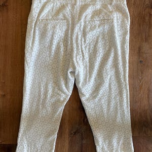May include: White capri trousers with an eyelet design. The trousers have a tailored fit with a button and zip closure, belt loops, and two back pockets. The fabric features a delicate, openwork pattern.