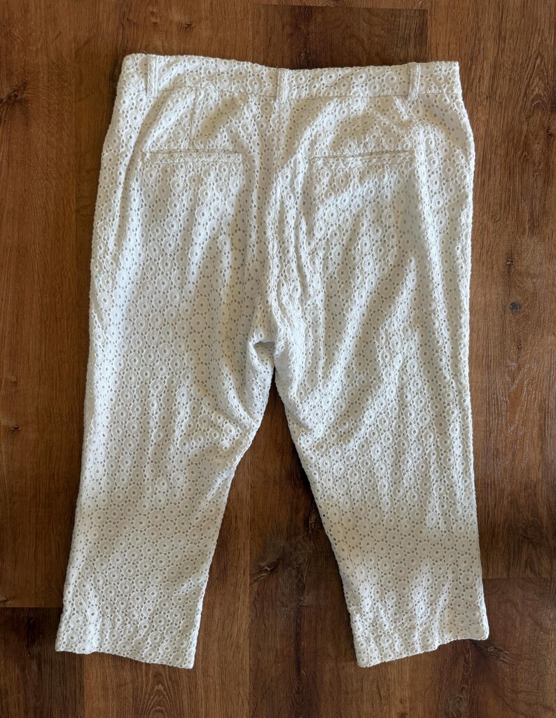 May include: White capri trousers with an eyelet design. The trousers have a tailored fit with a button and zip closure, belt loops, and two back pockets. The fabric features a delicate, openwork pattern.