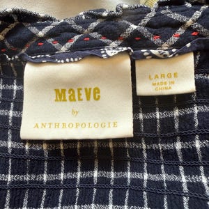 May include: Close-up of a navy blue and white checkered fabric. The fabric has a textured, smocked appearance. A tag reads "Maeve by Anthropologie" and another tag indicates the size is "LARGE" and "MADE IN CHINA".