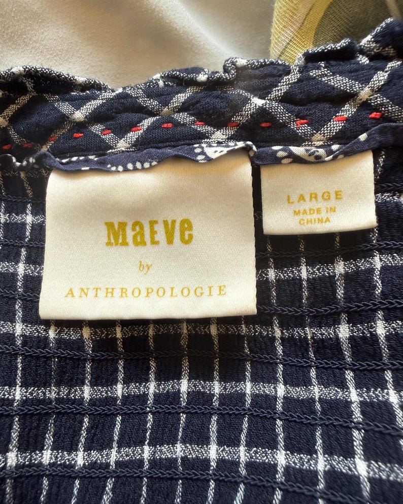 May include: Close-up of a navy blue and white checkered fabric. The fabric has a textured, smocked appearance. A tag reads "Maeve by Anthropologie" and another tag indicates the size is "LARGE" and "MADE IN CHINA".