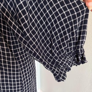 May include: Close-up of a navy blue dress with a white grid pattern. The sleeve features a ruffled cuff. The fabric appears to be soft and lightweight, suitable for warm weather. The dress is a classic style.