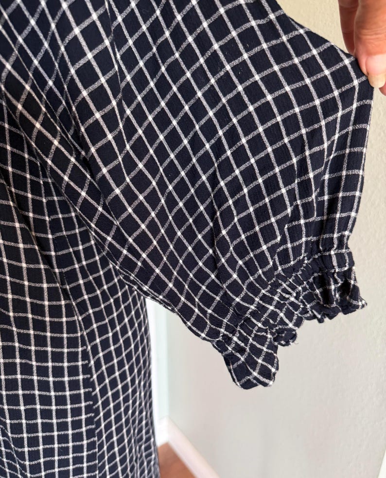 May include: Close-up of a navy blue dress with a white grid pattern. The sleeve features a ruffled cuff. The fabric appears to be soft and lightweight, suitable for warm weather. The dress is a classic style.