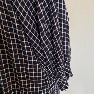 May include: Close-up of a navy blue and white checkered pattern fabric. The fabric appears to be part of a long-sleeved garment, with a gathered cuff visible. The pattern is a grid of white lines on a navy blue background.