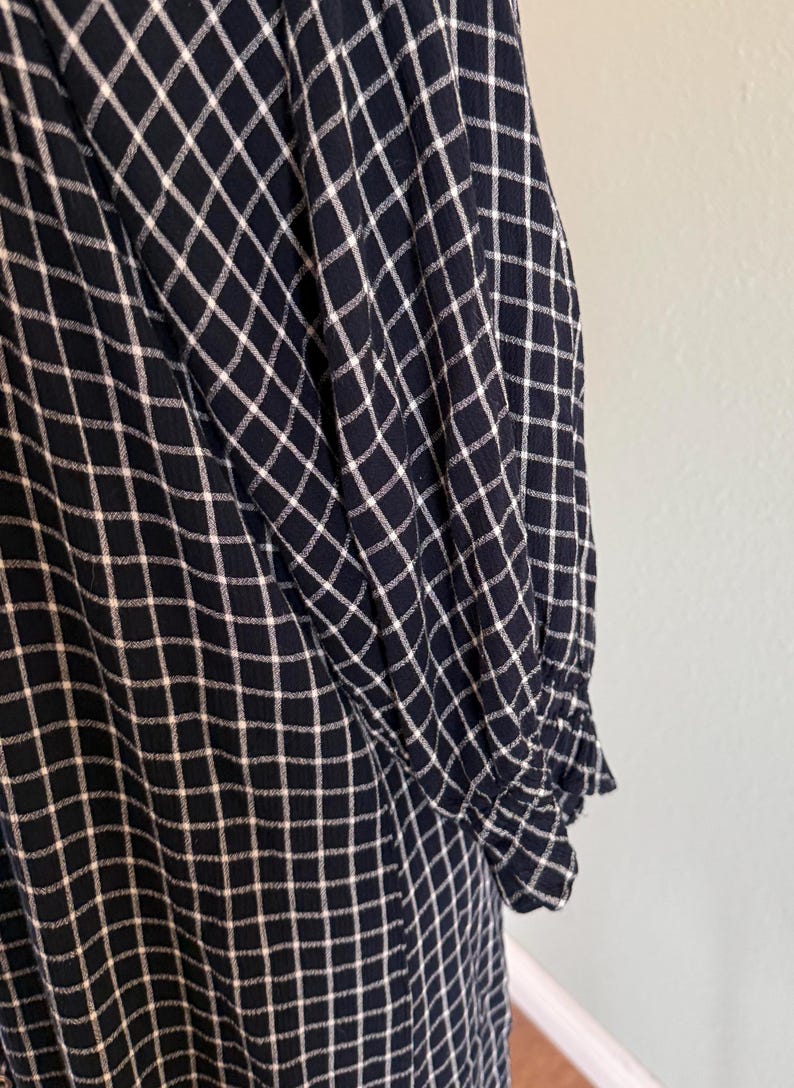 May include: Close-up of a navy blue and white checkered pattern fabric. The fabric appears to be part of a long-sleeved garment, with a gathered cuff visible. The pattern is a grid of white lines on a navy blue background.