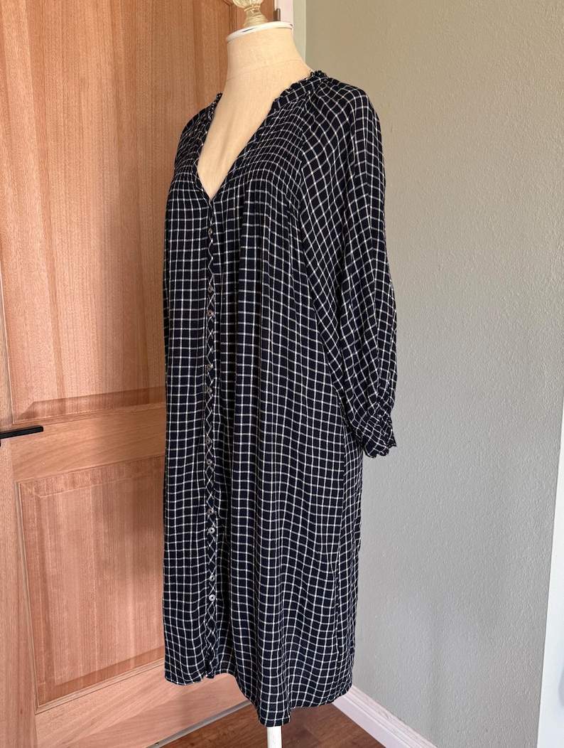 May include: A long-sleeved, knee-length dress in a navy blue and white grid pattern. The dress features a V-neckline, button-down front, and gathered detailing at the shoulders and cuffs. The dress is displayed on a mannequin.
