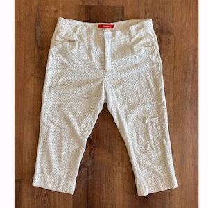 May include: Cream-coloured capri trousers featuring an intricate eyelet design. The capris have a button and zip fastening, belt loops, and pockets. The fabric looks lightweight, ideal for warmer weather.
