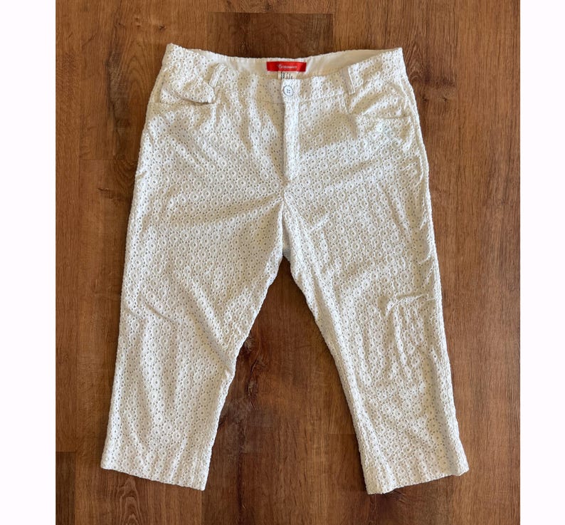 May include: Cream-coloured capri trousers featuring an intricate eyelet design. The capris have a button and zip fastening, belt loops, and pockets. The fabric looks lightweight, ideal for warmer weather.