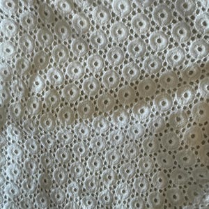 May include: Close-up of white eyelet fabric. The fabric has a repeating pattern of small circles, each with a central hole, creating a delicate, textured surface. The eyelet design is evenly spaced across the material, giving it a light and airy appearance.