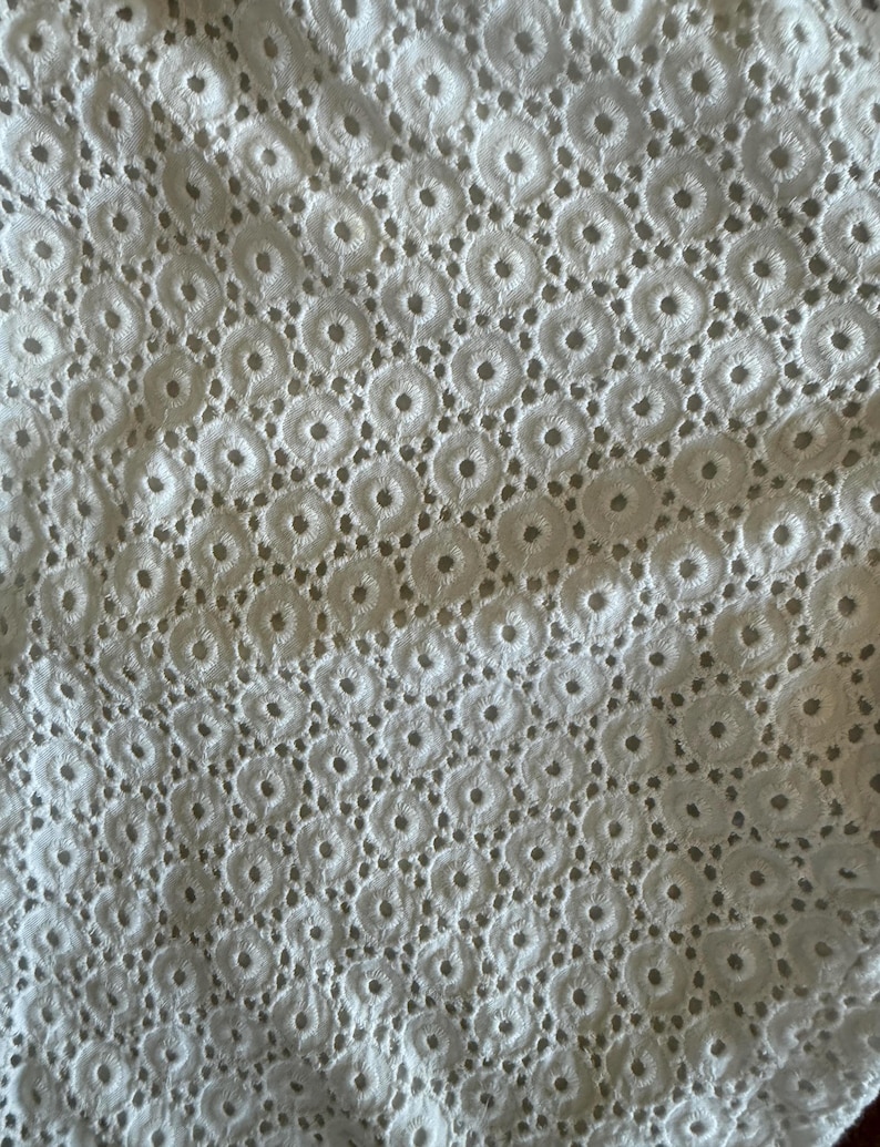 May include: Close-up of white eyelet fabric. The fabric has a repeating pattern of small circles, each with a central hole, creating a delicate, textured surface. The eyelet design is evenly spaced across the material, giving it a light and airy appearance.