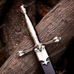 May include: A silver-toned metal dagger featuring a pointed pommel, a smooth grip, and a crossguard with decorative flared ends. The blade is partially encased in a black scabbard with a silver metal throat piece featuring an engraved design.