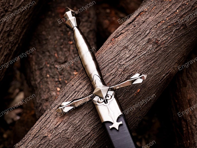 May include: A silver-toned metal dagger featuring a pointed pommel, a smooth grip, and a crossguard with decorative flared ends. The blade is partially encased in a black scabbard with a silver metal throat piece featuring an engraved design.