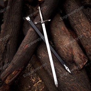 May include: A silver-toned metal sword with a detailed crossguard and pommel, positioned diagonally across a black scabbard. The scabbard features silver-colored metal fittings at the top and pointed tip, contrasting with the polished blade.