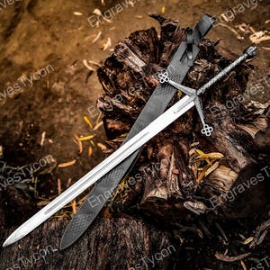 May include: A silver and black sword with a decorative handle and a long, sharp blade rests on a weathered wooden surface. A black sheath with a textured pattern lies beside the sword. The sword features ornate detailing.
