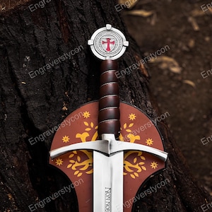 May include: A medieval-style sword with a silver blade and a brown leather-wrapped handle. The hilt features a decorative shield with a red cross emblem. The sword rests on a brown shield-shaped base with gold leaf designs. The text "NON NOB" is visible on the blade.