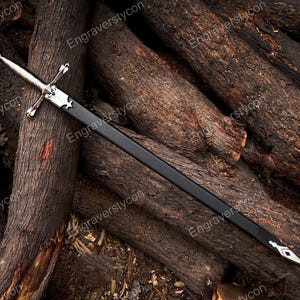 May include: A silver-toned metal sword with a cross-shaped guard and a black scabbard. The scabbard features a silver-colored metal fitting at the top near the hilt and a pointed silver-colored metal chape at the bottom tip of the blade casing.