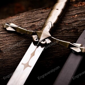 May include: A metallic sword featuring a polished blade with a central cutout design, a crossguard with pointed arms, and a textured handle. Beside the sword lies a black scabbard with a silver-toned metal tip, showcasing intricate geometric detailing.