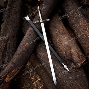 May include: A silver-toned metal sword with a detailed crossguard and pommel, positioned diagonally across a black scabbard. The scabbard features silver-colored metal fittings at the top and pointed tip, contrasting with the polished blade.