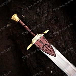 May include: A sword with a metallic silver blade and a dark reddish-brown hilt. The crossguard is gold-coloured and features the engraved text ANAKLYSMOS. A decorative reddish-brown triangular plate with a trident emblem is attached to the blade.