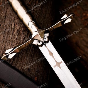 May include: A silver-toned metal sword featuring a detailed crossguard with pointed, faceted ends and a smooth, cylindrical handle. The blade displays a central decorative cutout pattern, and a black scabbard lies partially visible alongside it.
