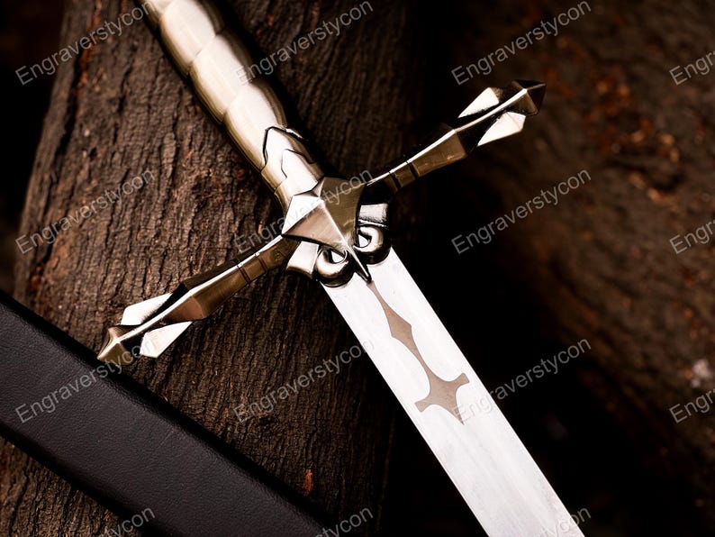 May include: A silver-toned metal sword featuring a detailed crossguard with pointed, faceted ends and a smooth, cylindrical handle. The blade displays a central decorative cutout pattern, and a black scabbard lies partially visible alongside it.