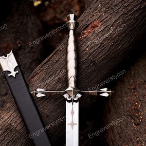 May include: A silver-toned metal sword featuring a ridged handle, a cross-shaped guard with pointed ends, and a blade with a decorative cutout near the hilt. A black scabbard with a silver metal throat piece is positioned alongside the sword.