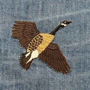 May include: Embroidered patch featuring a Canada goose in flight. The goose has brown wings, a tan body, and a black head with a white patch. The patch is on a blue denim background, showcasing the detailed stitching.