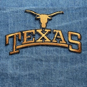 May include: Embroidered patch featuring the word "TEXAS" in a curved, gold-colored font, with a gold longhorn symbol above. The patch is set against a blue denim background, showcasing a classic, collegiate design.