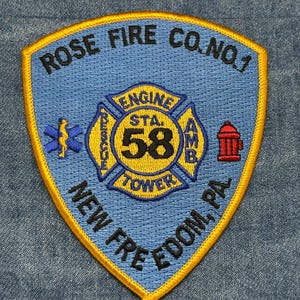 May include: A blue and gold embroidered patch for Rose Fire Co. No. 1, New Freedom, PA. The patch features a Maltese cross with the text "ENGINE STA. 58 TOWER" and other emergency service symbols. The patch is on a denim background.