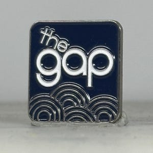 May include: A square pin featuring the text "the gap" in white, outlined in silver, set against a navy blue background. The pin has a silver border and a wave-like design at the bottom.