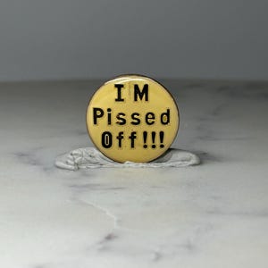 May include: A round, yellow pin with the text "I'M PISSED OFF!!!" in black lettering. The pin is set on a silver-colored, wax-like base. The background is a light, neutral color.