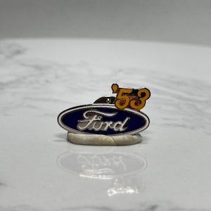 May include: A vintage Ford pin with a blue oval logo and the word "Ford" in white script. Above the logo is a gold-colored "'53" emblem. The pin is set on a light-colored base.