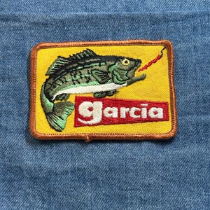 May include: Embroidered patch featuring a green and black fish with a red lure in its mouth, set against a yellow background. The patch has a brown border and the name "Garcia" in white letters on a red rectangle.