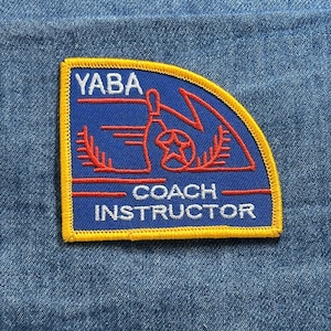 May include: A blue and yellow embroidered patch with the text "YABA COACH INSTRUCTOR". The patch features a bowling pin, a star, and other bowling-related graphics in red and white. The patch is set against a denim background.