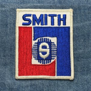 May include: Embroidered patch with the word "SMITH" in blue block letters above a graphic of the number "9" inside a blue and red square. The patch has a white border and is set against a denim background.