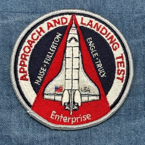 May include: Embroidered patch featuring a white space shuttle design with red and blue accents. The patch reads "Approach and Landing Test" and "Enterprise". The patch includes the text "Haise-Fullerton" and "Engle-Truly".