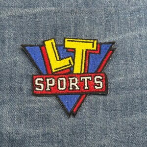 May include: Embroidered patch with the text "LT SPORTS" in a retro design. The patch features a blue triangle with red accents, yellow lettering, and a red rectangle with white text.