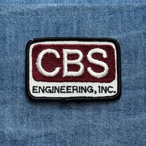 May include: A rectangular patch with the letters "CBS" in white, embroidered on a maroon background. The patch is outlined in black and white, with the words "ENGINEERING, INC." below the letters. The patch is set against a blue denim background.