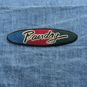 May include: An embroidered patch with the word "Bandag" in white script. The patch is oval-shaped with a blue, burgundy, and green color scheme. The patch is set against a denim background.