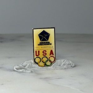 May include: A rectangular pin featuring the Olympic rings and the text "USA." The pin includes the text "1992 Albertville" and a logo. The pin has a gold border and a cream-colored background.