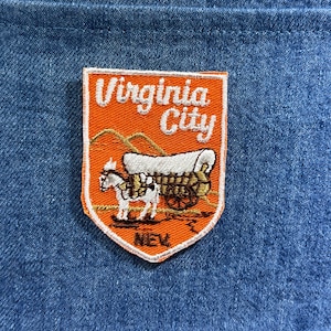 Puede incluir: Parche bordado con el texto "Virginia City" sobre un fondo naranja. El parche representa una carreta cubierta tirada por un caballo. La palabra "NEV" está en la parte inferior, todo sobre un fondo de mezclilla azul.