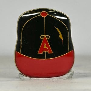 May include: A baseball cap-shaped pin with a black top and red brim. The pin features a gold outline and a red letter "A" on the front. The pin is likely a collectible or souvenir.