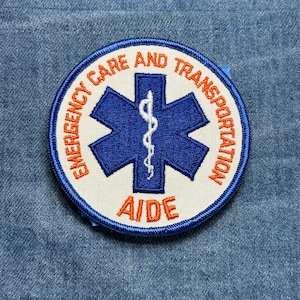 May include: Embroidered patch with the words "EMERGENCY CARE AND TRANSPORTATION" in orange around a white circle. Inside is a blue star of life symbol with a white medical staff and the word "AIDE" in orange.