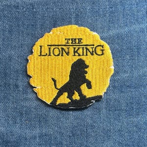 May include: Embroidered patch featuring the words "THE LION KING" above a black silhouette of a lion. The patch has a yellow background and a black border. The patch is on a blue denim background.
