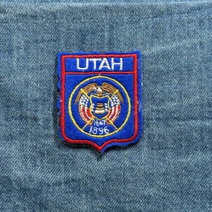 May include: A blue and red embroidered patch featuring the word "UTAH" at the top. Below the word is a circular emblem with the state seal, including the dates 1847 and 1896. The patch is on a blue denim background.